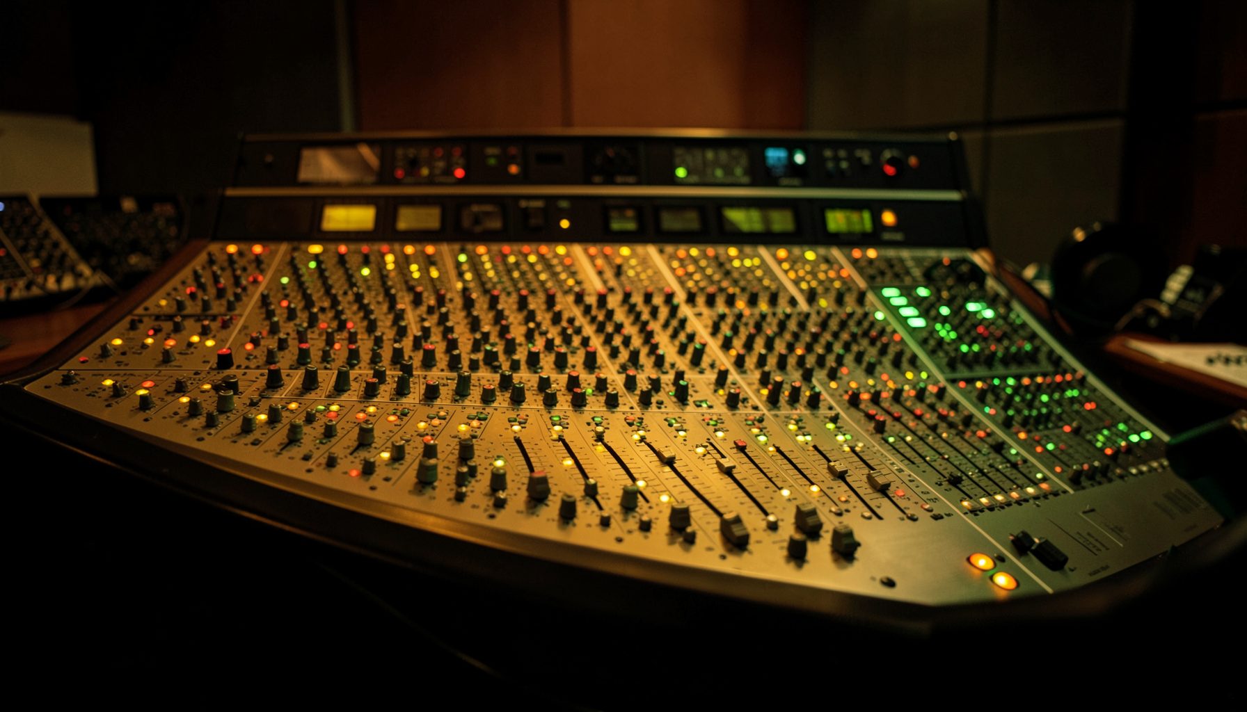 Large professional mixing console with rows of knobs, faders, and illuminated meters in a dimly lit studio control room, warm amber and green glow