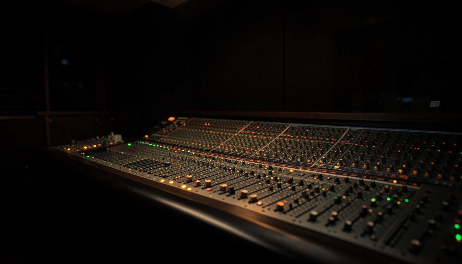 Dark studio mixing console with rows of illuminated red, green, and amber buttons and faders, moody low-light scene