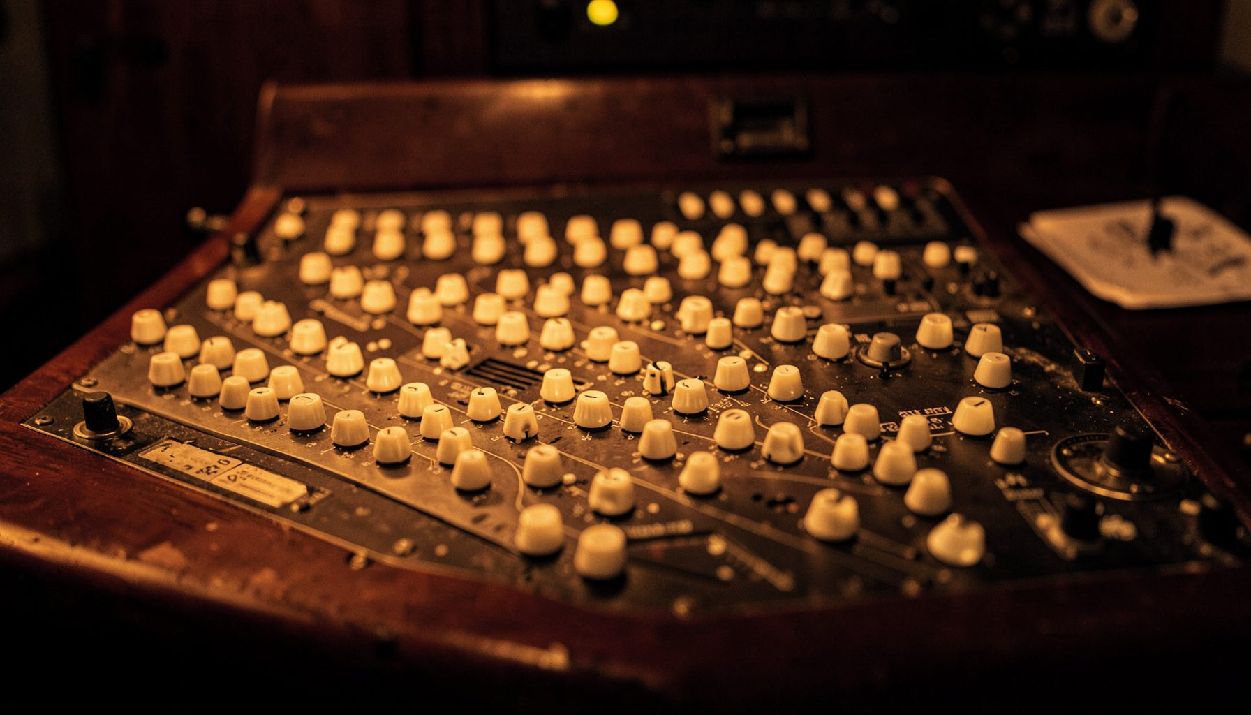 Warmly lit vintage mixing console with rows of cream-colored knobs and dark paneling, rich amber glow across the control surface